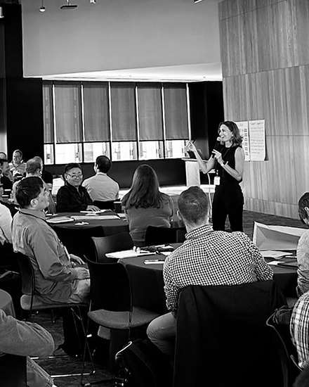 A full room of participants at round tables, with Annemarie Boss standing at the front beside a flip chart.