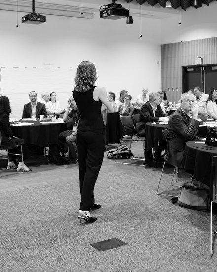 Annemarie Boss with her back to the camera, facing several dozen participants seated at round tables.