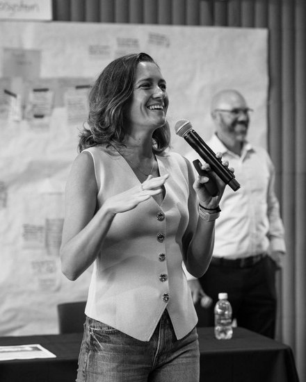 Annemarie Boss speaking into a handheld microphone in front of a wall of sticky notes, a colleague standing behind her.
